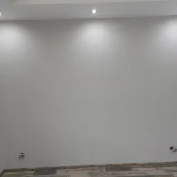 Wnętrze świeżo wykończonego pomieszczenia z gładkimi, szarymi ścianami, drewnopodobną podłogą i zamontowanymi punktowymi lampami sufitowymi, widoczne puszki elektryczne w ścianach przed montażem...