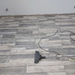 Świeżo położona podłoga z paneli imitujących drewno w odcieniach szarości, częściowo odkurzona, widoczny odkurzacz i kabel na tle jasnych ścian i sufitu z wystającymi puszkami elektrycznymi.