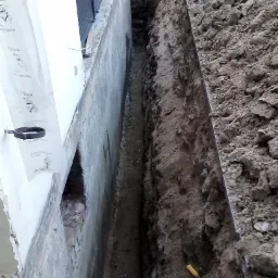 Wykop wąskoprzestrzenny wzdłuż fundamentu budynku, widoczna folia ochronna przymocowana do ściany, łopatka leżąca na ziemi.