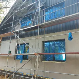 Dom w trakcie prac elewacyjnych: rusztowanie, okna zabezpieczone folią, nałożony tynk surowy, widoczna drewniana boazeria na górnej części elewacji.