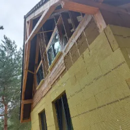 Budynek w trakcie ocieplania, widoczna warstwa izolacji z wełny mineralnej Rockwool na elewacji, drewniana konstrukcja dachu i okna z czarnymi ramami, w tle sosnowy las.