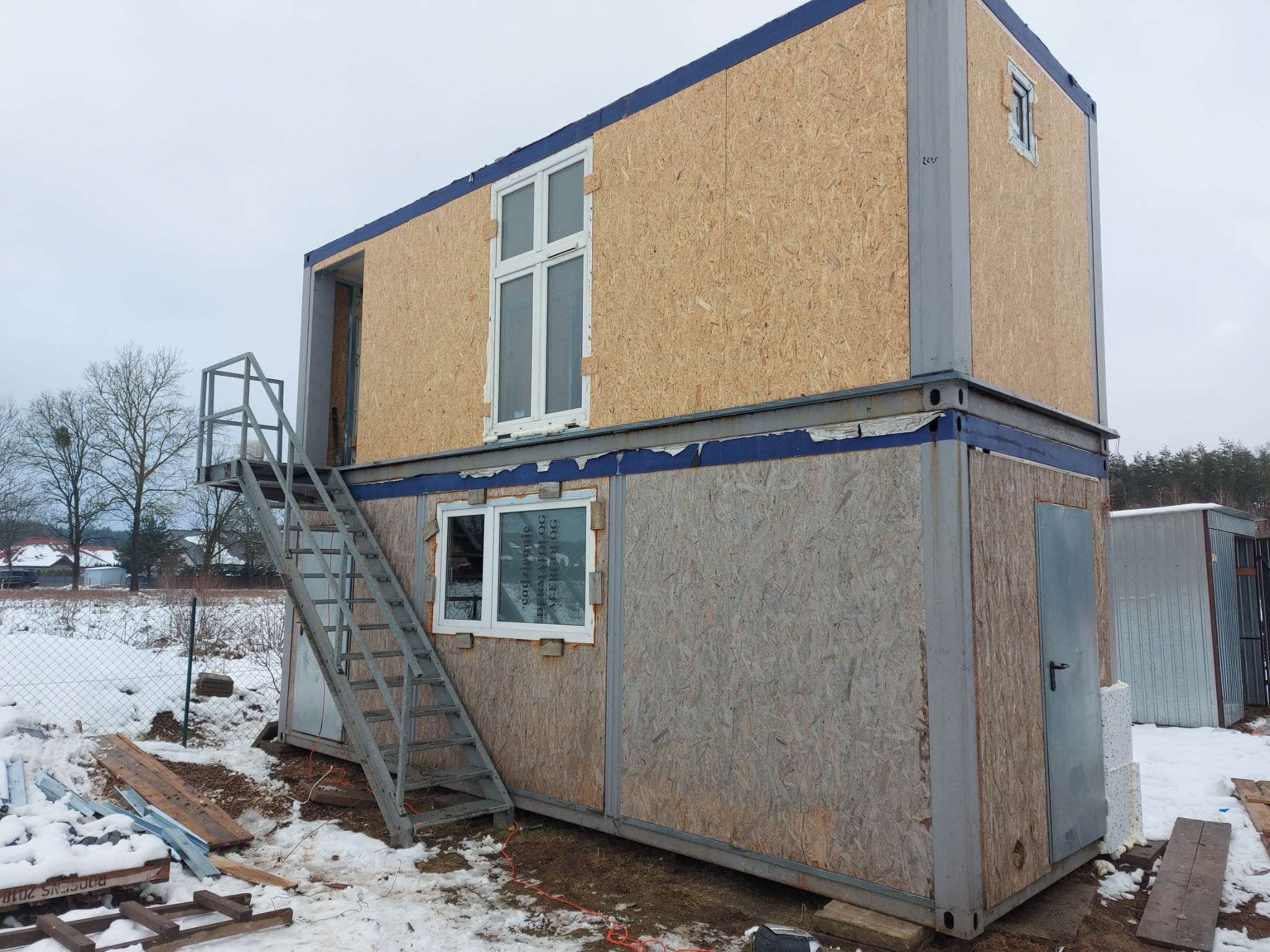 Dwu-kondygnacyjny budynek modułowy z płyty OSB w trakcie montażu, z metalowymi schodami zewnętrznymi i oknami PCV, na zimowym placu budowy.