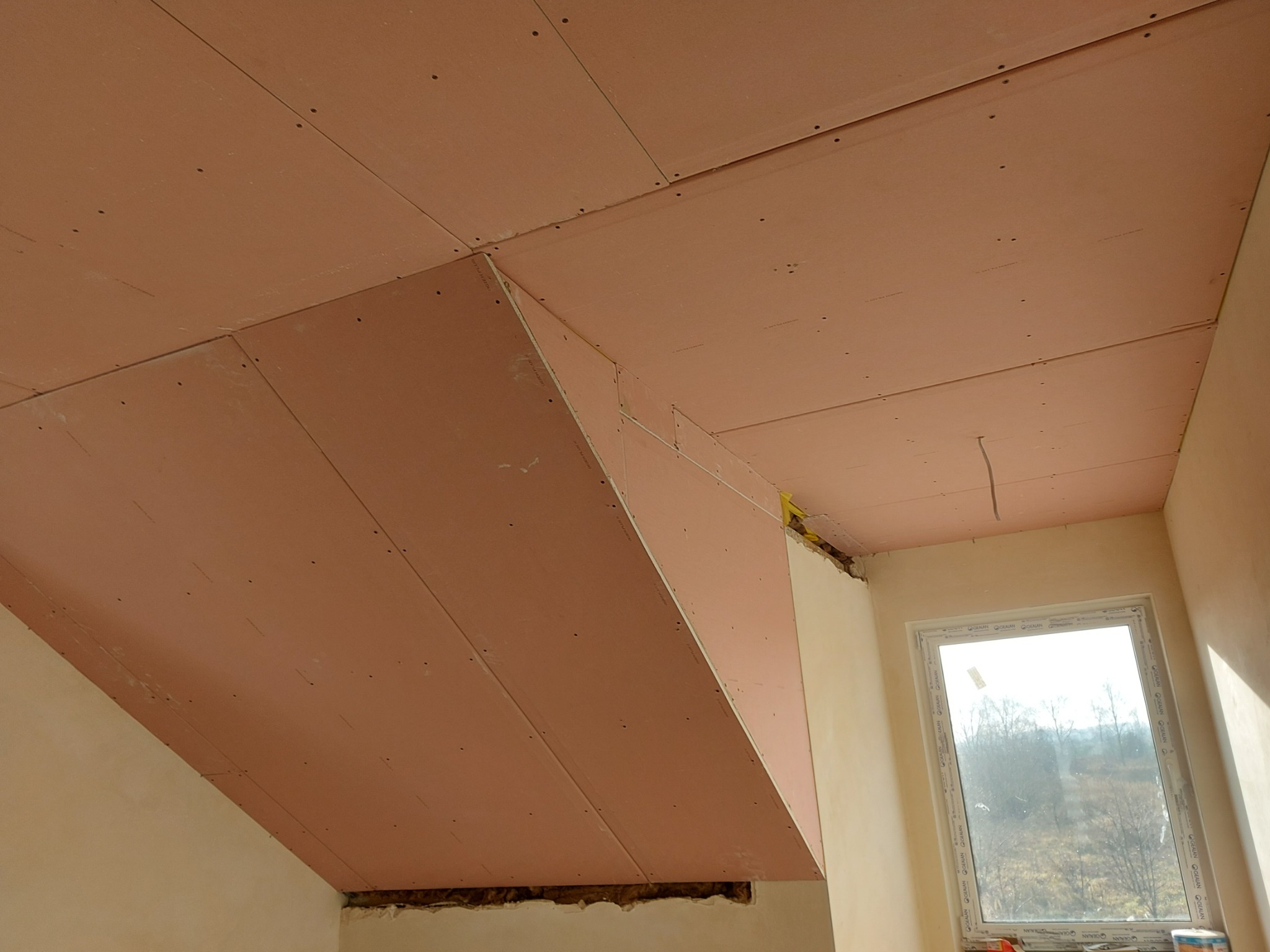 Montaż różowych płyt kartonowo-gipsowych na skośnym poddaszu, widoczne wkręty i łączenia płyt, okno w tle