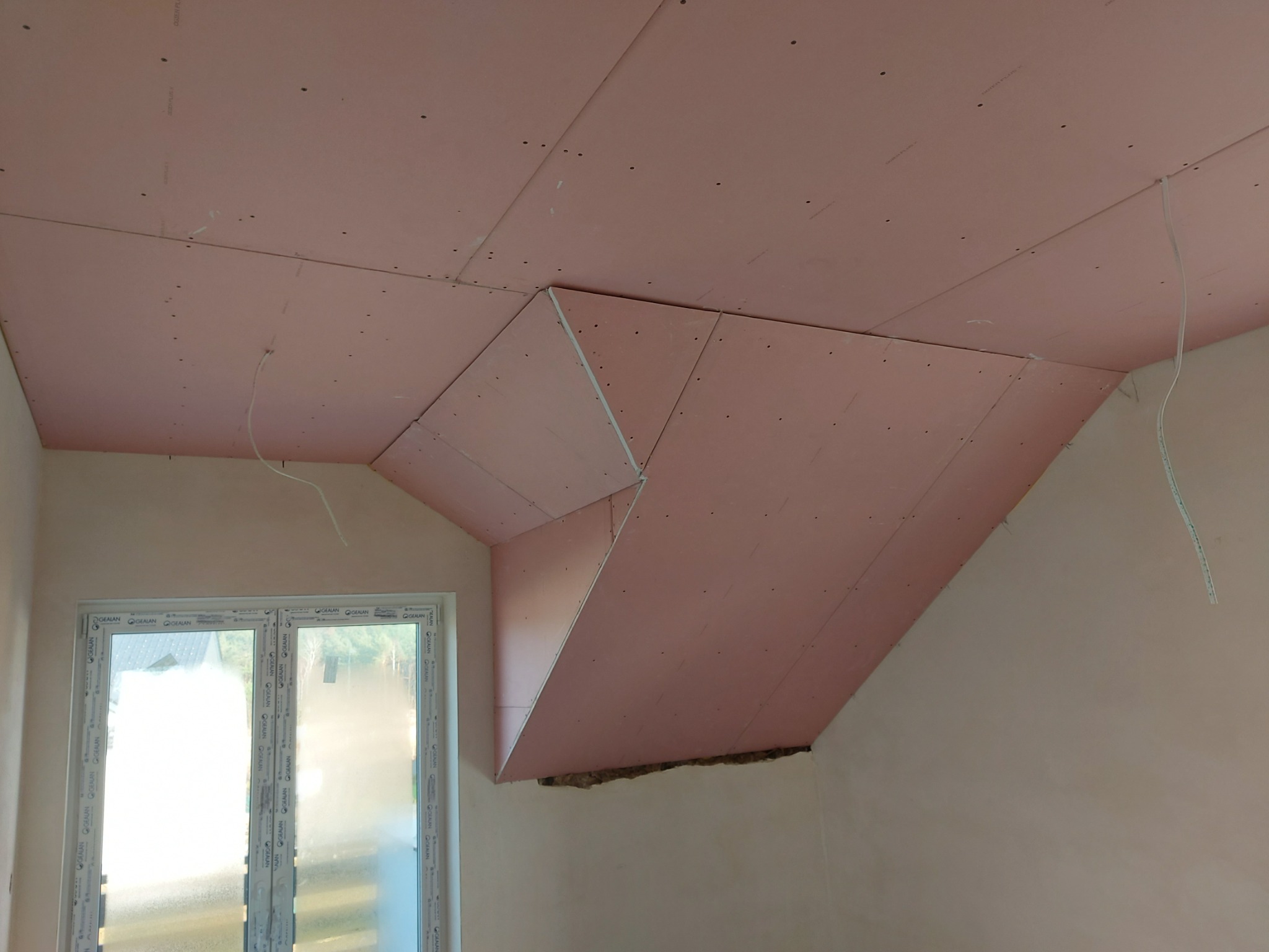 Montaż różowych płyt gipsowo-kartonowych na skośnym poddaszu, widoczne łączenia i niedokończona izolacja przy ścianie, okno z PCV w tle.
