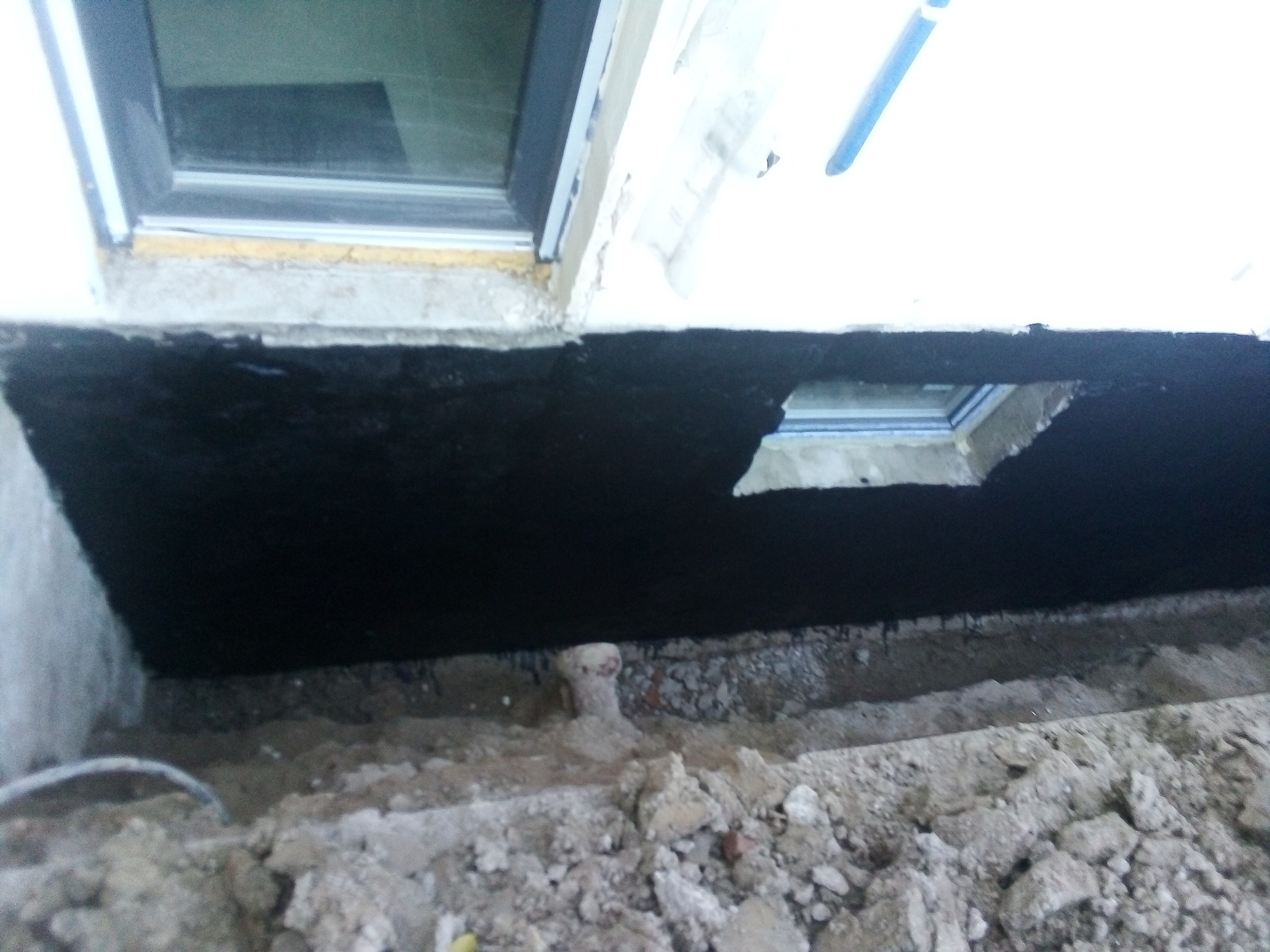 Czarna izolacja przeciwwilgociowa fundamentu domu, widoczne okno piwniczne i grunt.