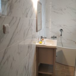 Piotr Dabrowski - Wykończona łazienka z marmurowymi płytkami na ścianach, umywalką z szafką w kolorze taupe i drewnopodobną podłogą, widok na wannę ze słuchawką prysznicową.