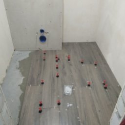 Piotr Dabrowski - Układanie płytek podłogowych imitujących drewno w łazience, z użyciem klipsów poziomujących i krzyżyków dystansowych; widoczny fragment ściany z instalacją podtynkową WC.