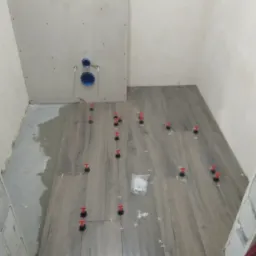 Układanie płytek podłogowych imitujących drewno w łazience, z użyciem klipsów poziomujących i krzyżyków dystansowych; widoczny fragment ściany z instalacją podtynkową WC.