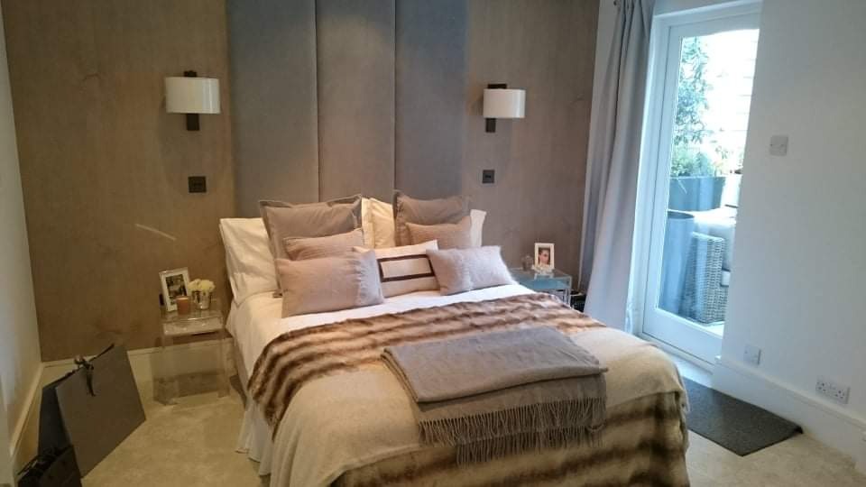 Elegancka sypialnia z łóżkiem w odcieniach beżu i szarości, ściany wykończone w dwóch odcieniach z kinkietami, szklane stoliki nocne z dekoracjami, widok na balkon przez przeszklone drzwi.