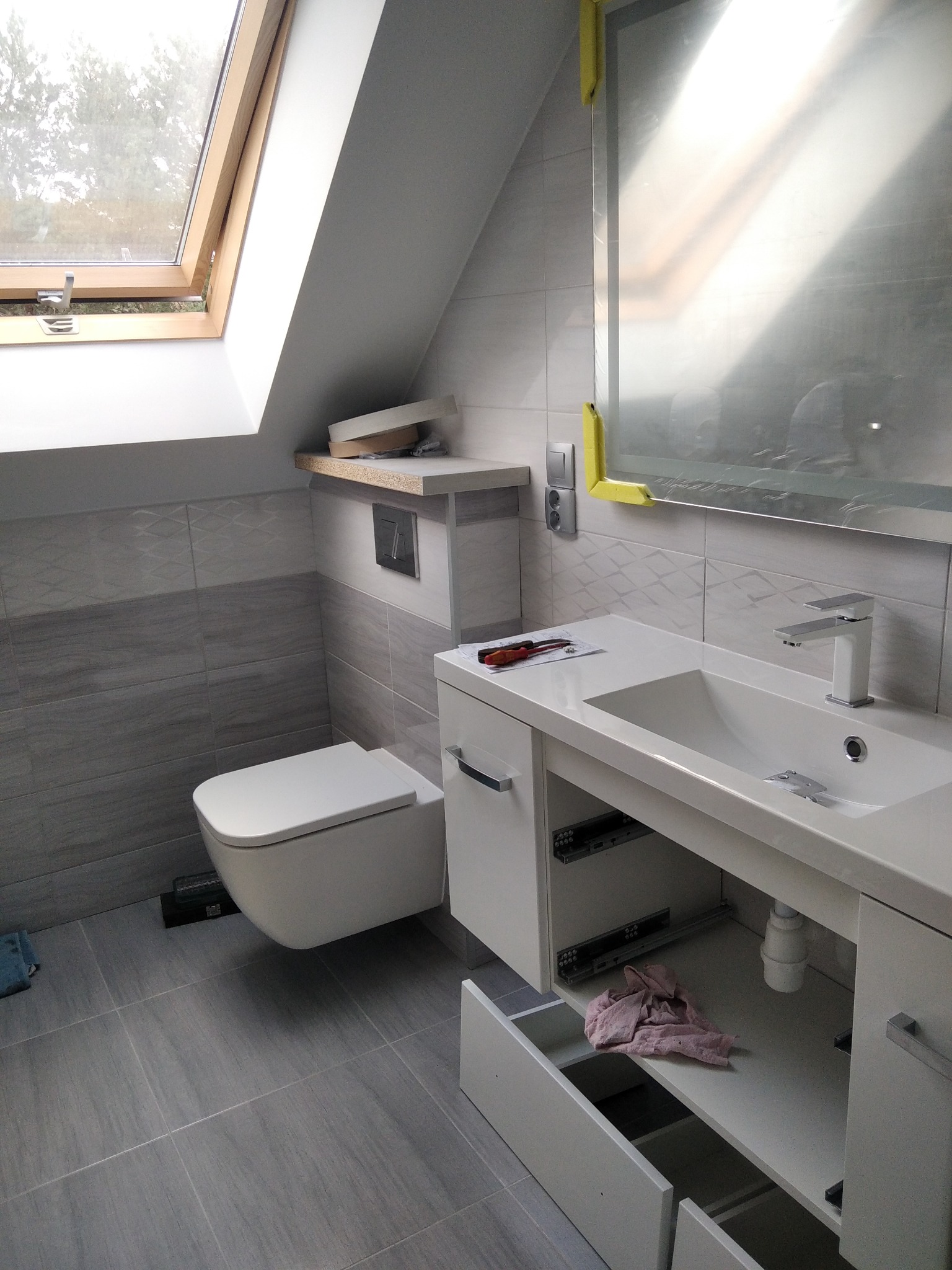 Remont łazienki w Nowej Soli: Ukończona aranżacja z szarymi płytkami, podwieszaną toaletą, białą szafką z umywalką i oknem dachowym. Widoczne drobne niedociągnięcia wykończeniowe, takie jak folia...