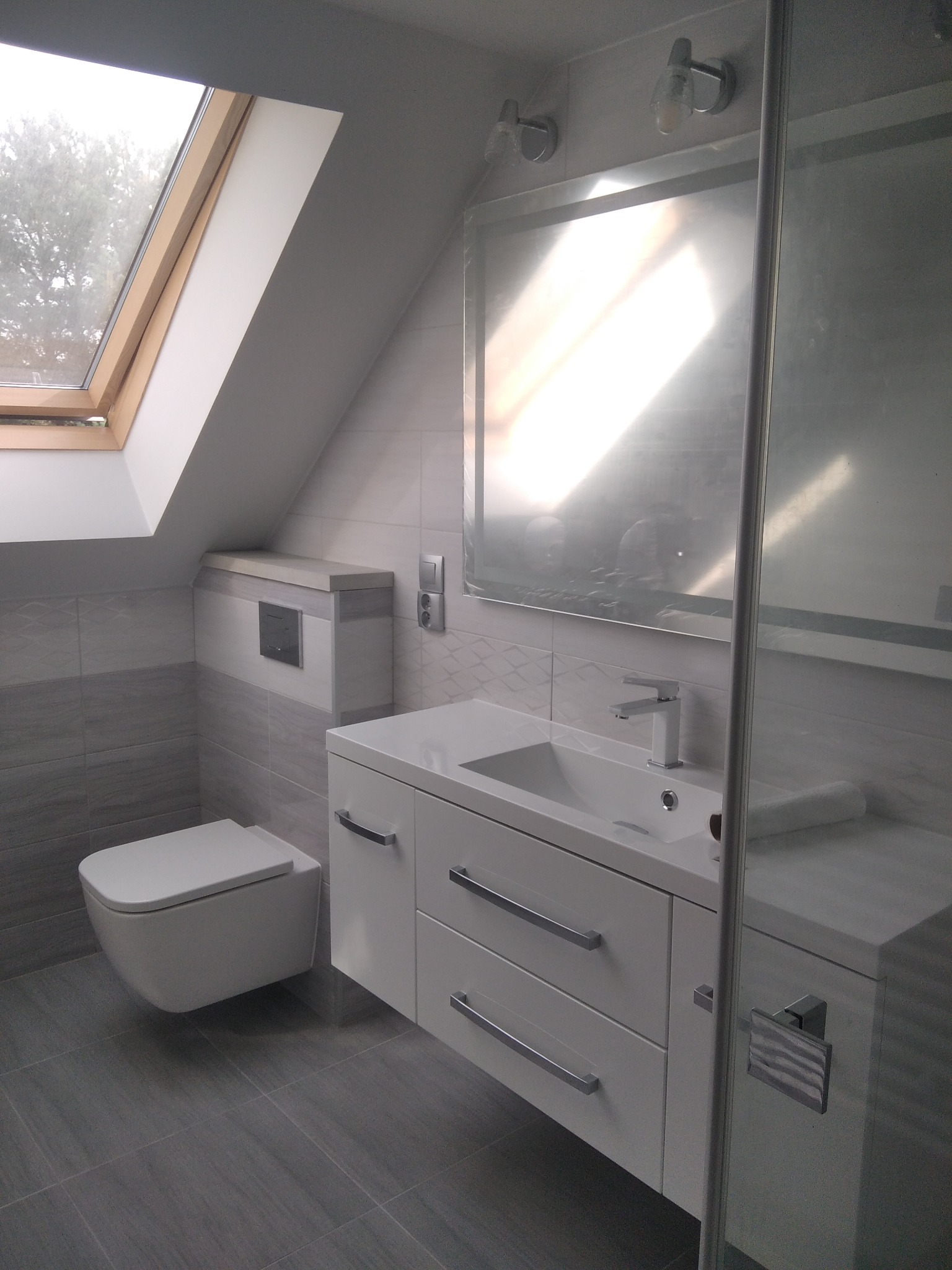 Nowoczesna łazienka na poddaszu z oknem dachowym, szare płytki imitujące drewno, biała wisząca toaleta i szafka z umywalką podwieszaną, widoczna część kabiny prysznicowej.