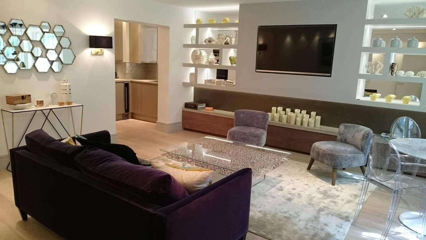 Elegancki salon z fioletową sofą, stolikiem kawowym z blatem lastryko, dekoracyjną ścianą z półkami podświetlanymi LED, oraz widocznym fragmentem kuchni w tle.