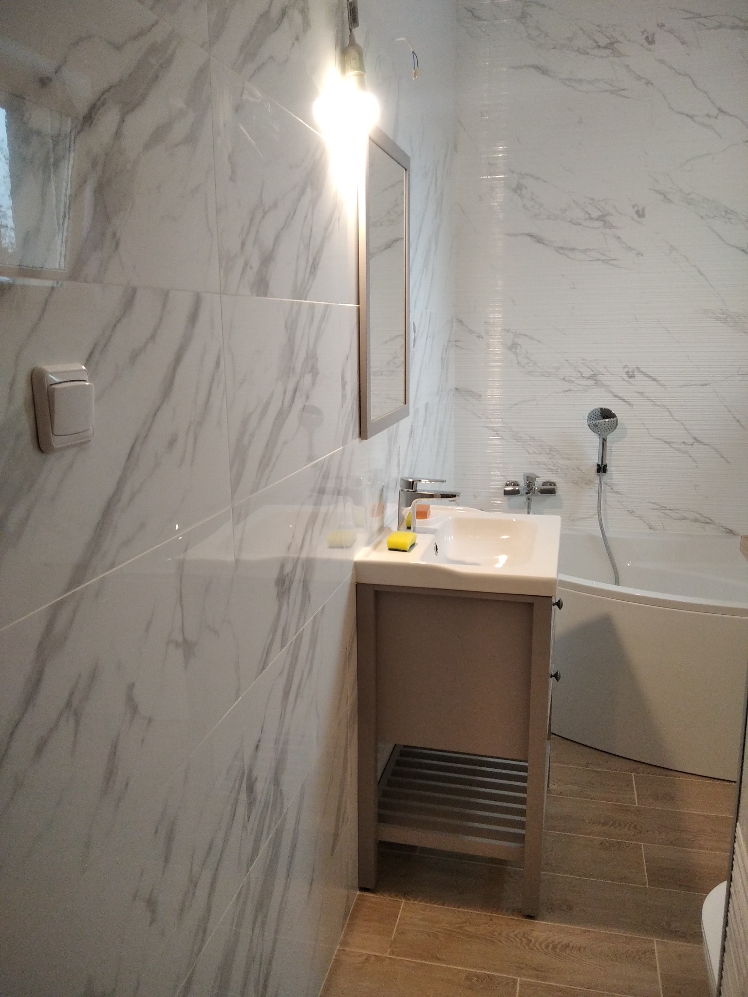 Wykończona łazienka z marmurowymi płytkami na ścianach, umywalką z szafką w kolorze taupe i drewnopodobną podłogą, widok na wannę ze słuchawką prysznicową.