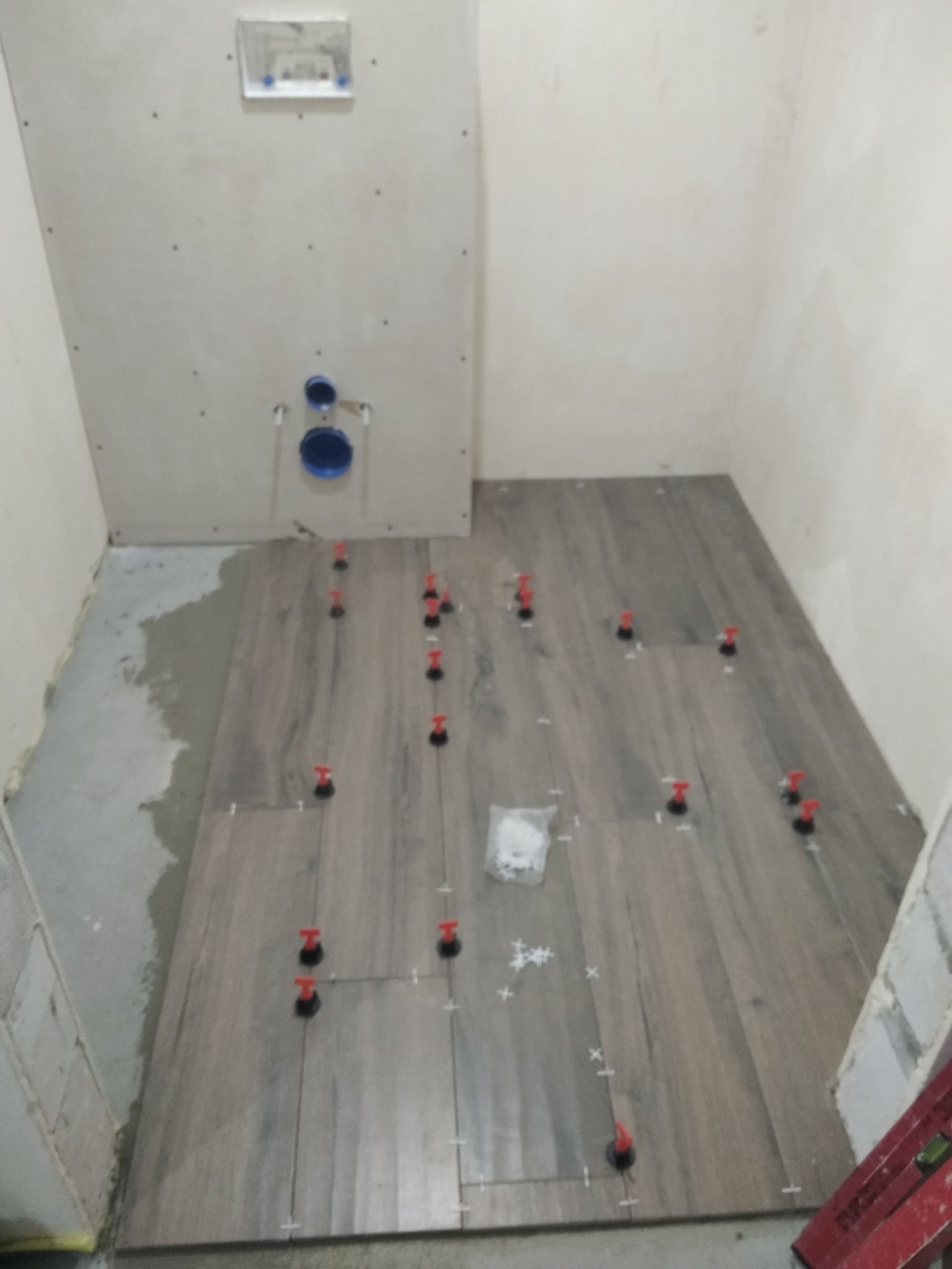 Układanie płytek podłogowych imitujących drewno w łazience, z użyciem klipsów poziomujących i krzyżyków dystansowych; widoczny fragment ściany z instalacją podtynkową WC.