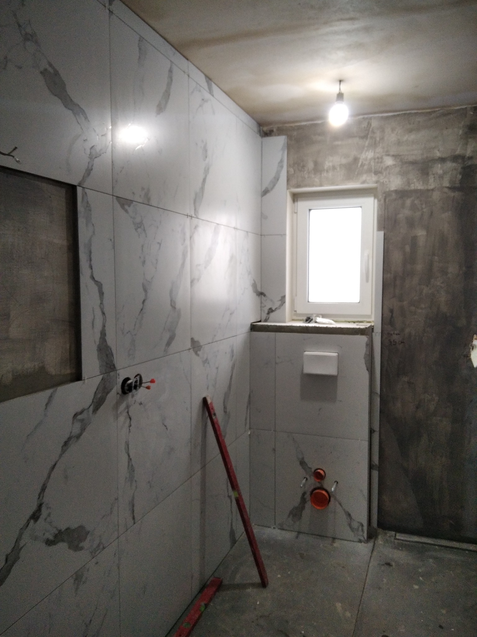 Łazienka w trakcie remontu: ściany wyłożone białymi płytkami imitującymi marmur, widoczne instalacje wodno-kanalizacyjne, fragment ściany przygotowany do położenia gładzi, poziomica oparta o ścianę.