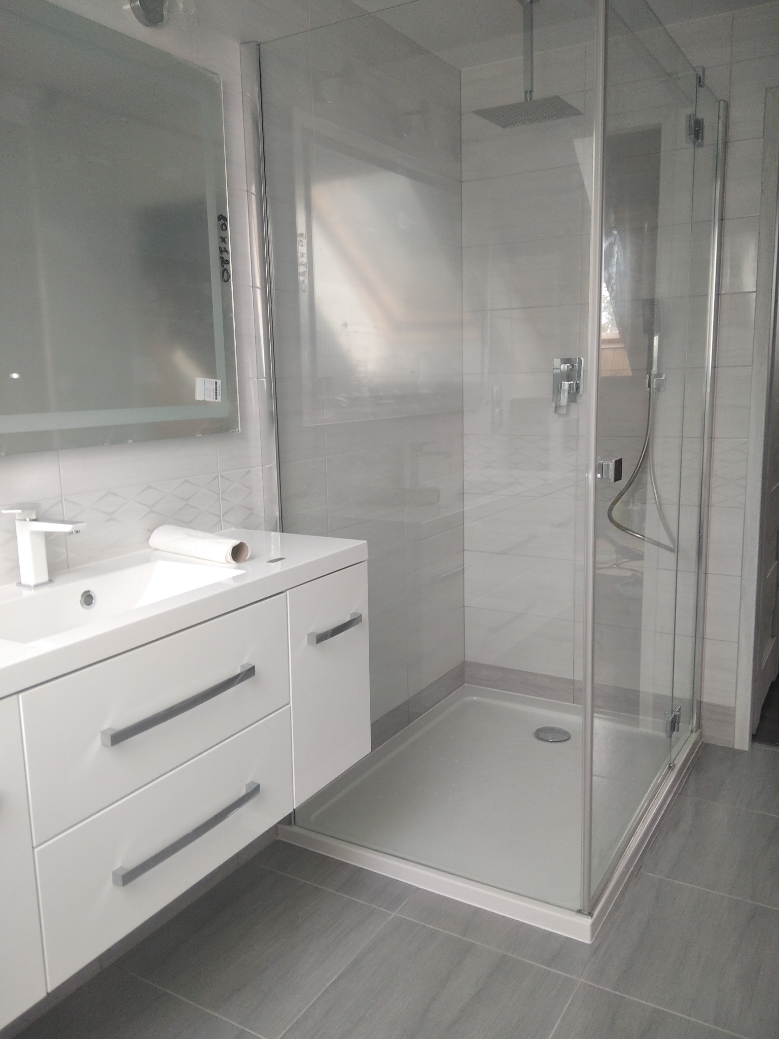 Nowoczesna łazienka z kabiną prysznicową typu walk-in, białą szafką z umywalką i lustrem z podświetleniem LED, wykończona szarymi płytkami.