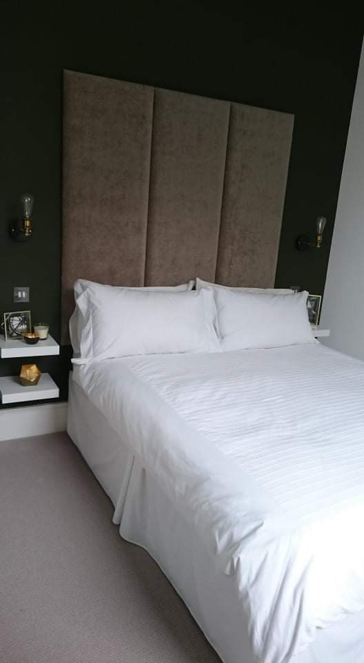 Sypialnia z wysokim, tapicerowanym wezgłowiem w odcieniach beżu, białą pościelą, ciemnozieloną ścianą i minimalistycznymi półkami nocnymi z dekoracjami.