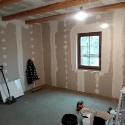 Wnętrze w trakcie prac wykończeniowych: surowe drewniane belki stropowe, ściany częściowo pokryte płytami gipsowo-kartonowymi i szpachlą, okno z drewnianą ramą, oraz narzędzia i materiały budowlane...