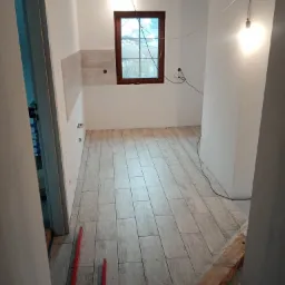 Układanie płytek podłogowych w jasnym pomieszczeniu z drewnianym oknem i surowymi belkami stropowymi, widoczne poziomice i fragmenty niezakończonej podłogi.