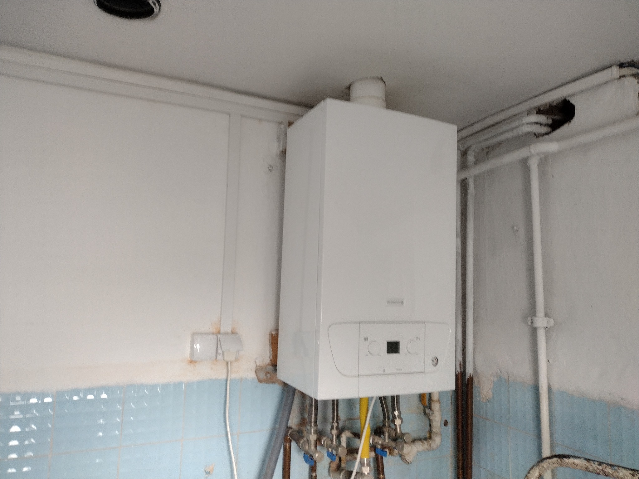 Biały piec gazowy kondensacyjny zamontowany na ścianie z niebieskimi kafelkami, widoczne rury i przyłącza wodne oraz gazowe.