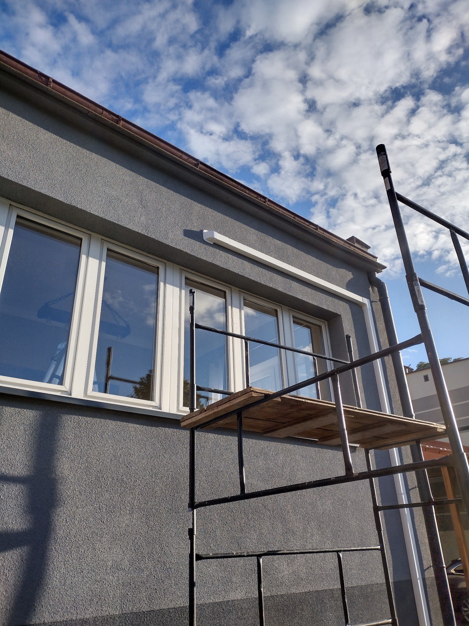 Szarego koloru elewacja domu z zamontowanymi oknami, na której widać rusztowanie z drewnianą platformą roboczą, a nad oknami zamontowany element instalacji.