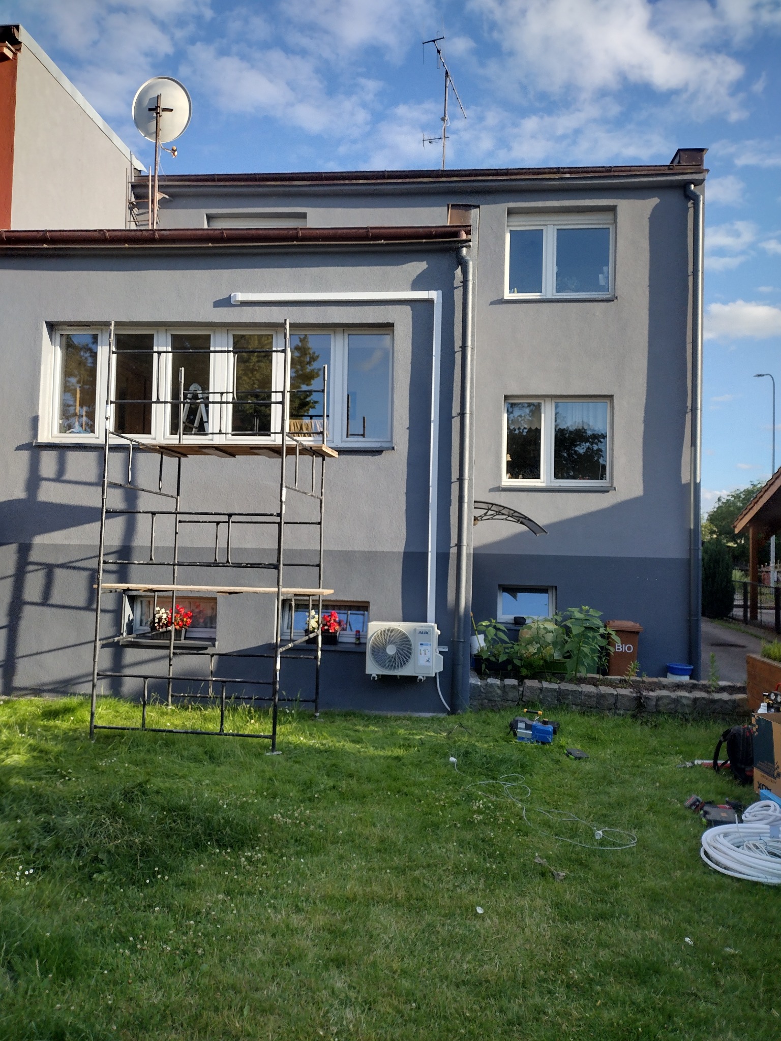 Szary, dwukondygnacyjny budynek w Sławnie z zamontowaną klimatyzacją zewnętrzną, widoczny z poziomu trawnika, z rozstawionym rusztowaniem i narzędziami do montażu na trawie.