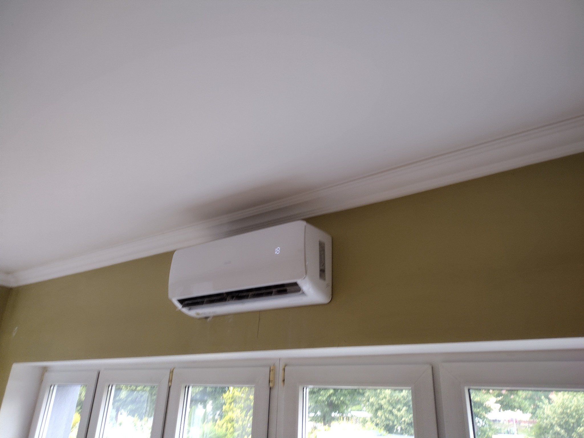 Biała klimatyzacja ścienna z wyświetlaczem temperatury '18' zamontowana nad oknem w pokoju z oliwkową ścianą i białym sufitem z dekoracyjną listwą.
