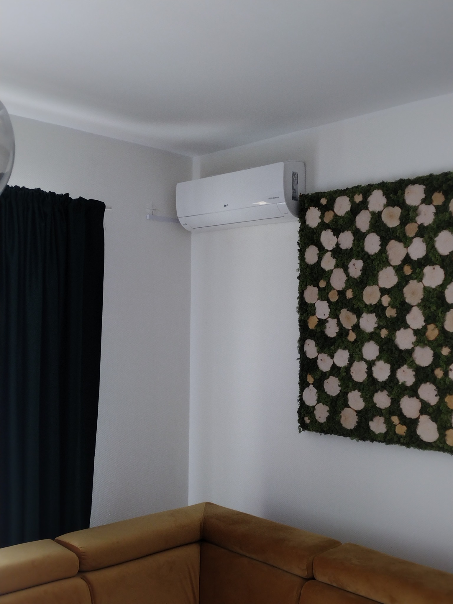 Biały klimatyzator LG zamontowany na ścianie w salonie, obok dekoracji z mchu i drewnianych plastrów, widoczny fragment sofy w kolorze musztardowym, czarne zasłony.