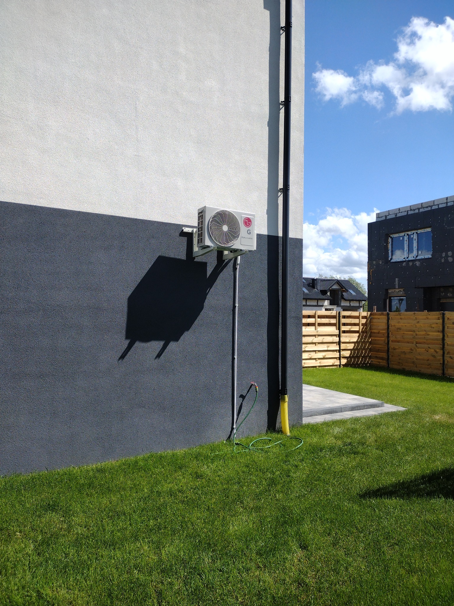 Zewnętrzna jednostka klimatyzacji LG na dwukolorowej elewacji domu z widocznym odprowadzeniem skroplin i fragmentem trawnika.
