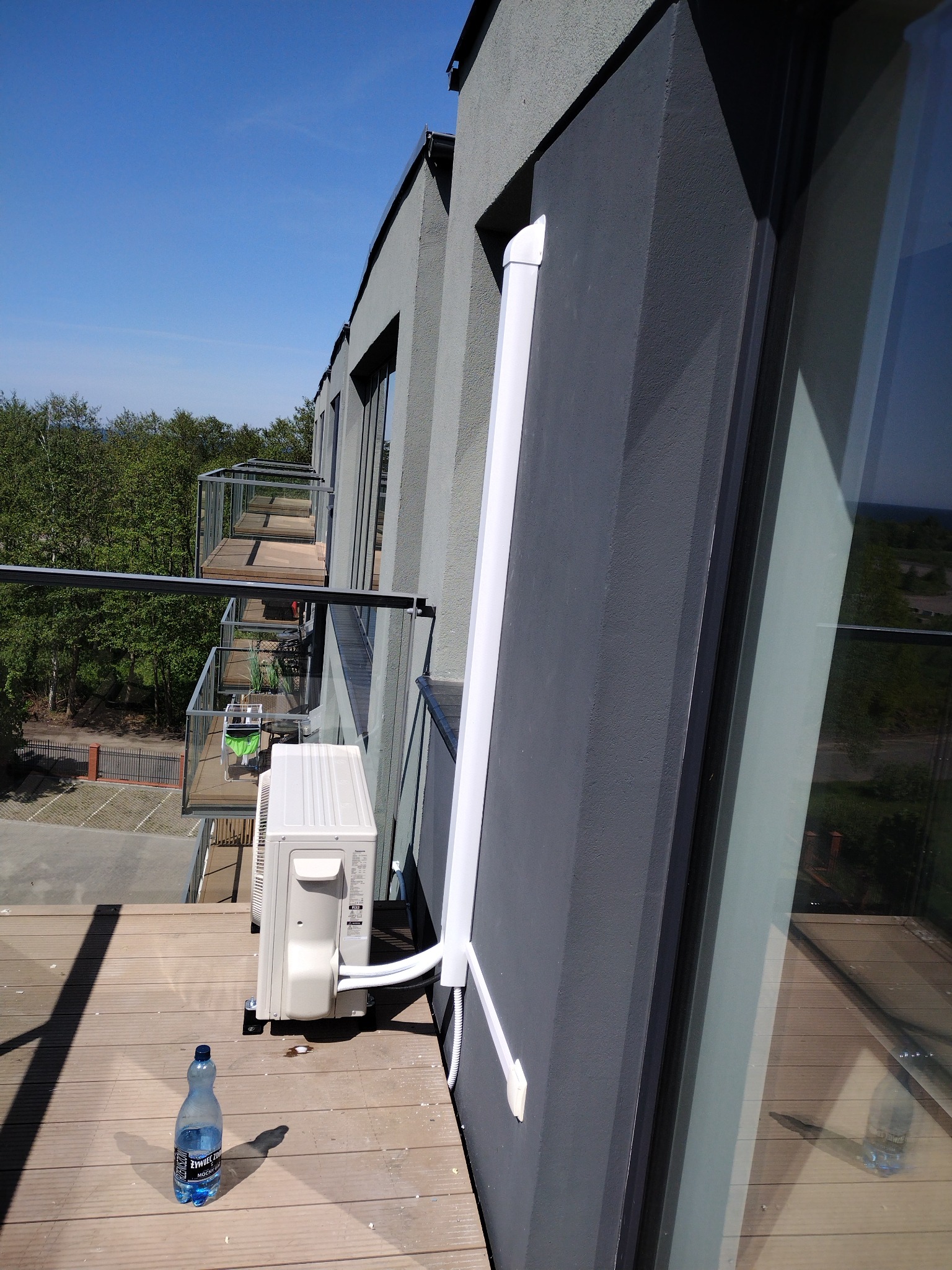 Zewnętrzna jednostka klimatyzacji zamontowana na balkonie nowoczesnego budynku, obok butelka wody mineralnej, widok z góry na sąsiednie balkony i drzewa.