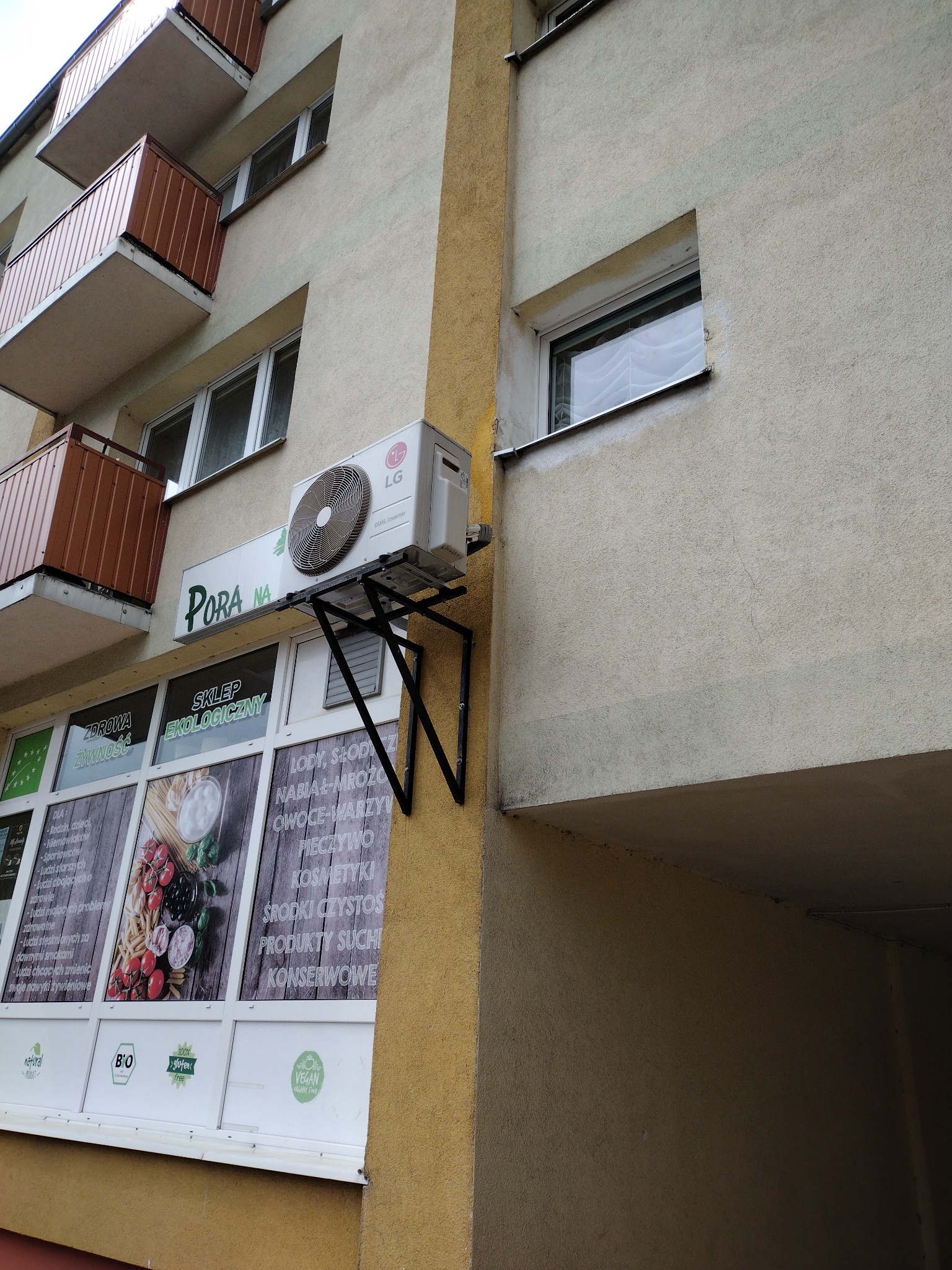Zewnętrzna jednostka klimatyzacji LG zamontowana na ścianie budynku nad witryną sklepu 'Pora na', widoczny wspornik mocujący, balkony powyżej i okno obok.