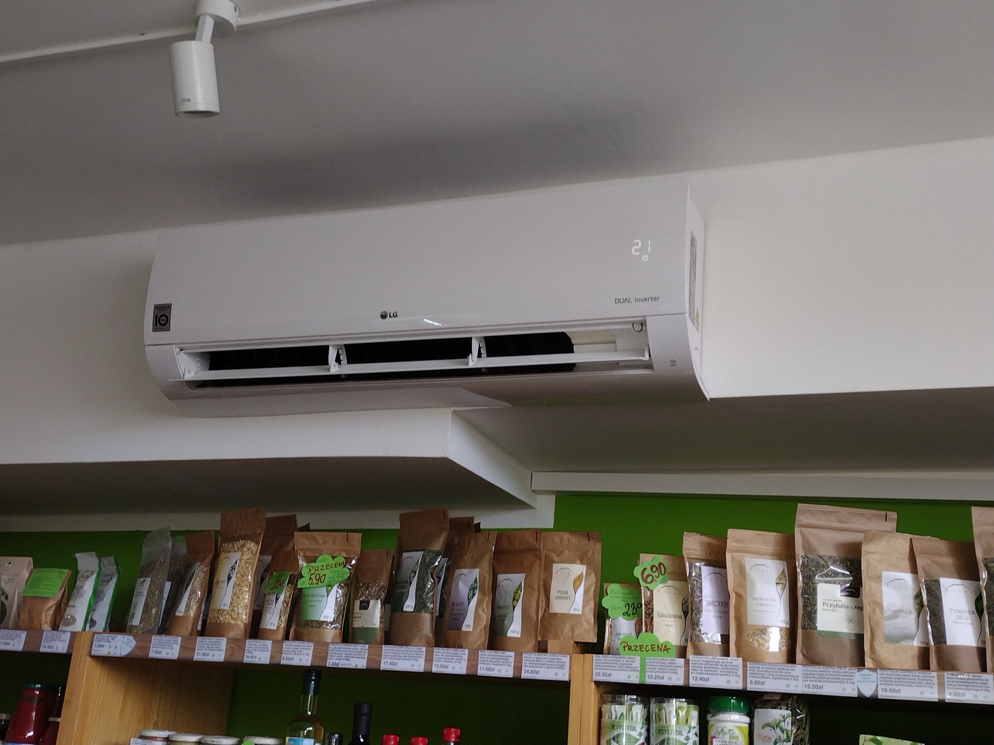 Biała klimatyzacja LG Dual Inverter zamontowana na ścianie w sklepie z ziołami i przyprawami, widoczna półka z produktami w ekologicznych opakowaniach.