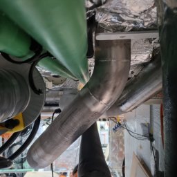 Piotr Nowak - Wnętrze techniczne z rurami metalowymi i elementami instalacji, widoczne przewody i izolacja. Układ rur w nietypowym ujęciu z dołu.