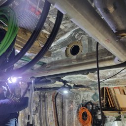 Piotr Nowak - Instalacja rur: pracownik w kombinezonie łączy rury w przestrzeni technicznej, widoczne przewody, izolacja i oświetlenie robocze.