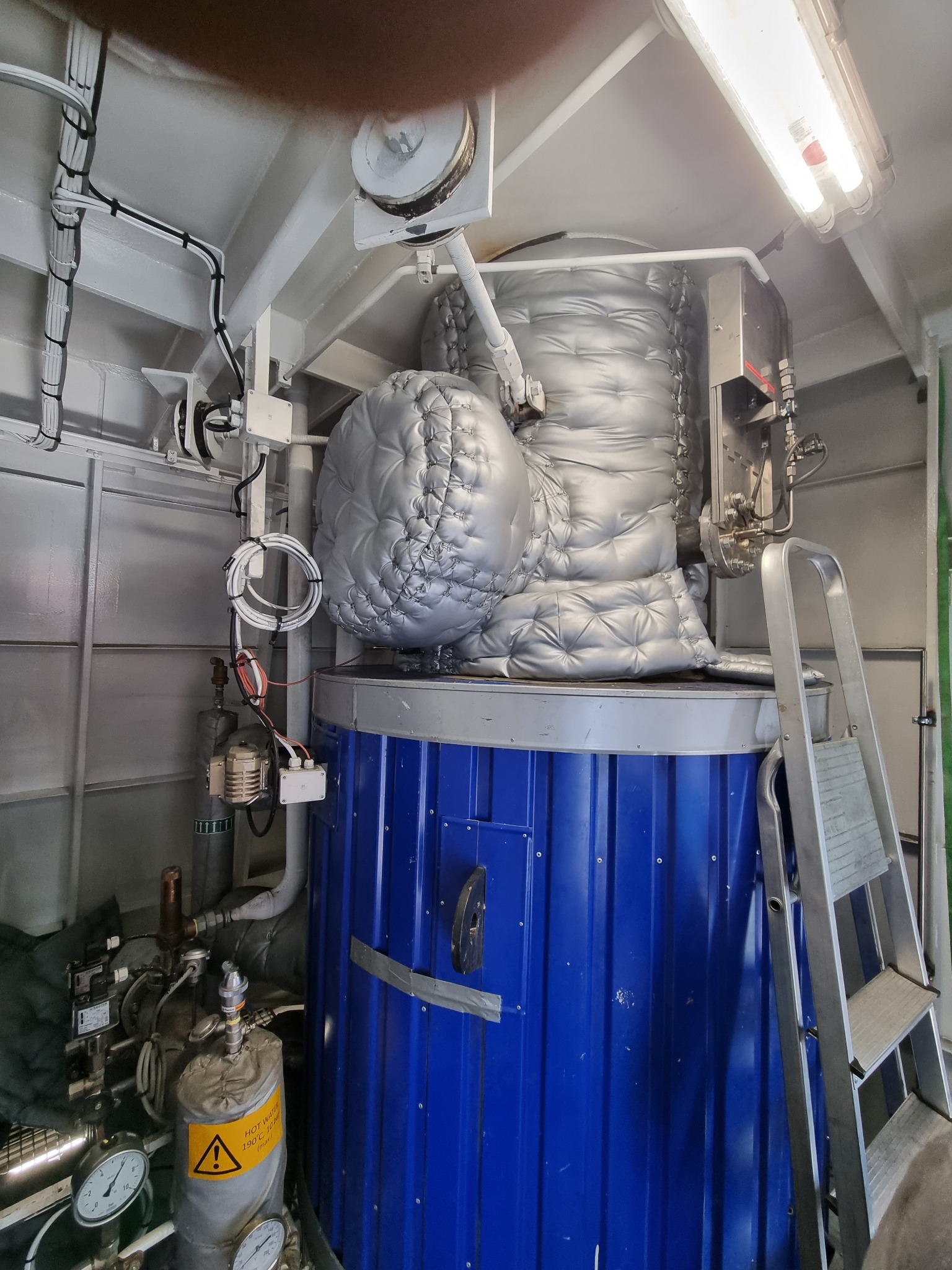Instalacja przemysłowa z niebieskim zbiornikiem i srebrną izolacją termiczną, widoczne rury, kable i drabina w hali o metalowych ścianach. Oznakowanie ostrzegawcze.