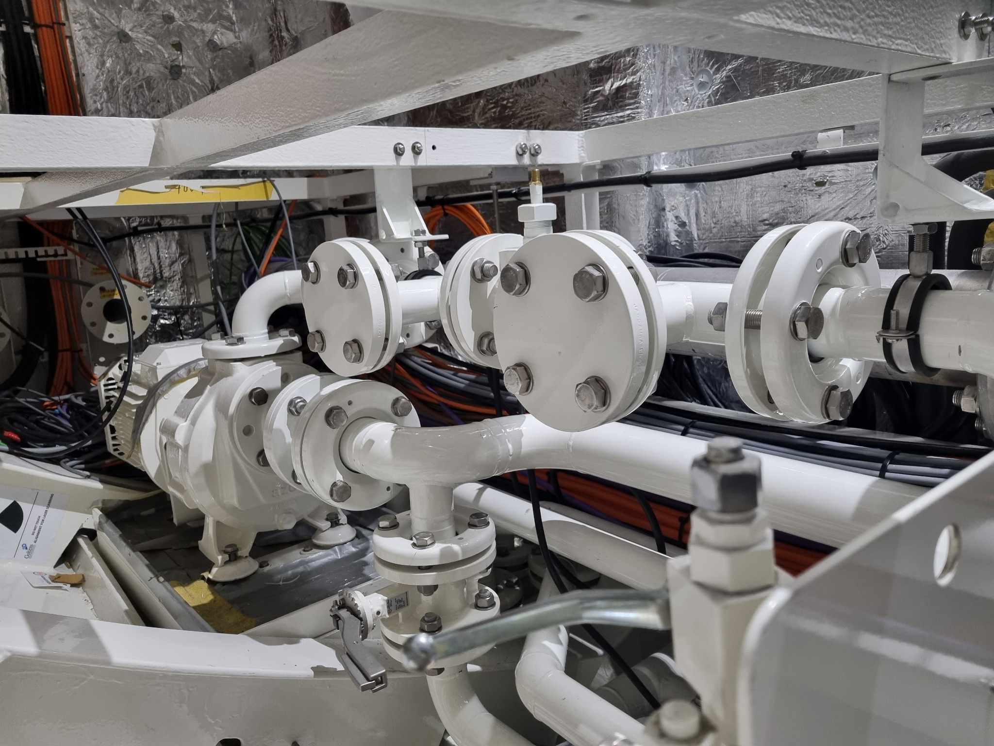 Biała instalacja rurowa z licznymi złączkami i mocowaniami, widoczne przewody elektryczne i elementy konstrukcyjne. Złożony system instalacji w przestrzeni technicznej.