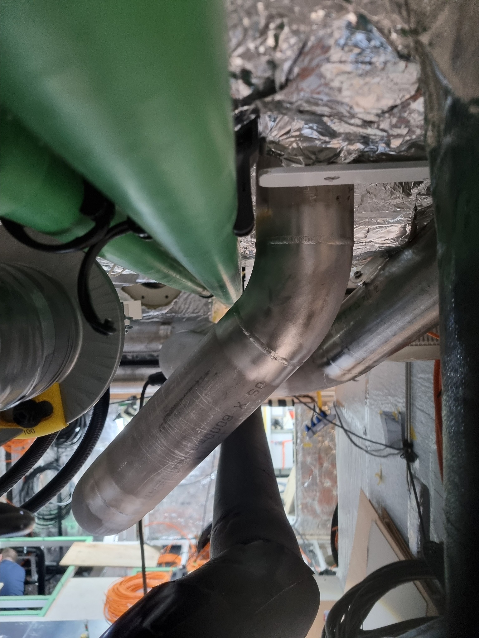 Wnętrze techniczne z rurami metalowymi i elementami instalacji, widoczne przewody i izolacja. Układ rur w nietypowym ujęciu z dołu.