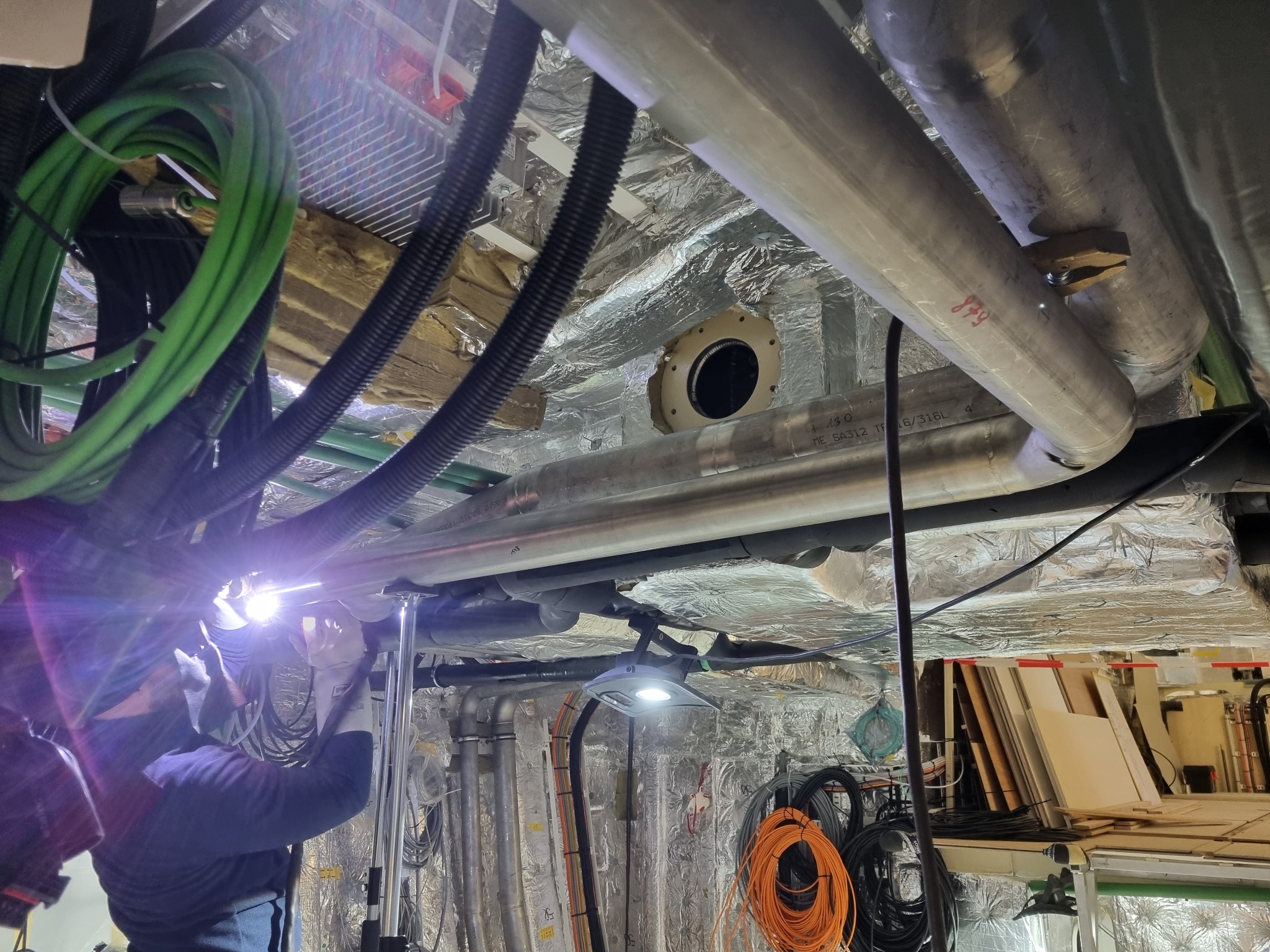 Instalacja rur: pracownik w kombinezonie łączy rury w przestrzeni technicznej, widoczne przewody, izolacja i oświetlenie robocze.