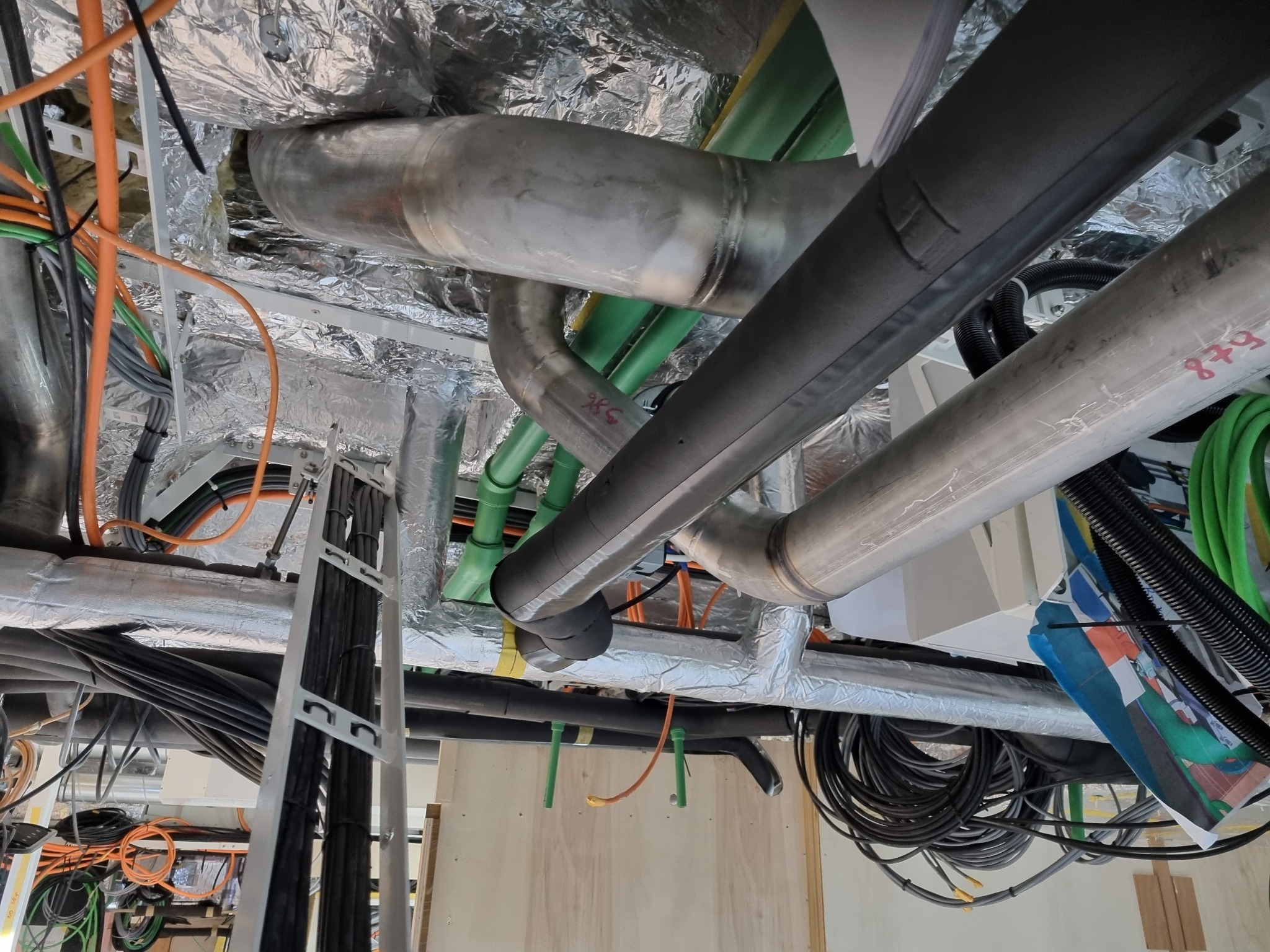 Skomplikowana instalacja rur i kabli w budynku, widoczne metalowe rury, izolacja, kable w różnych kolorach, srebrna folia izolacyjna na suficie. Ujęcie z dołu.