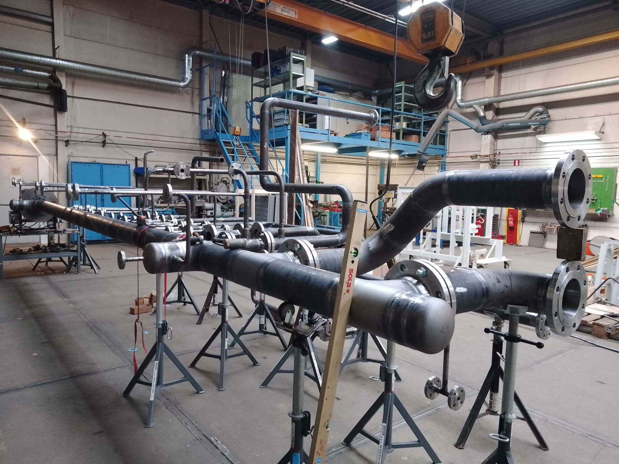 Wnętrze hali z fragmentem spawanej konstrukcji rurociągu na stojakach. Widoczny poziomica, suwnica i instalacje pod stropem.