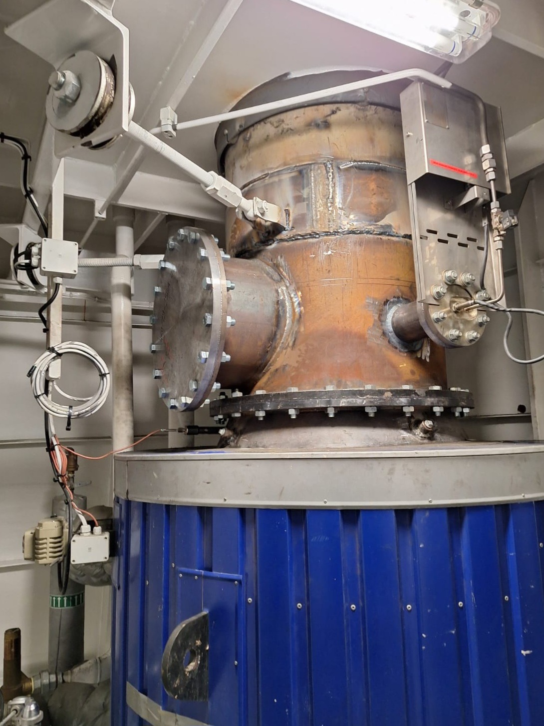 Metalowa konstrukcja zbiornika z widocznymi spawami i elementami instalacji, osadzona na niebieskiej, ryflowanej podstawie. Widoczne przewody i mocowania.