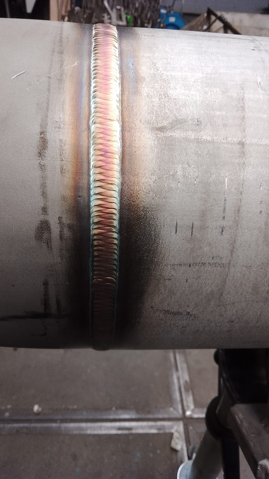 Spoina na rurze metalowej z widocznymi śladami ciepła i tęczowymi przebarwieniami, wykonana precyzyjnie, z bliska, na tle warsztatowym.