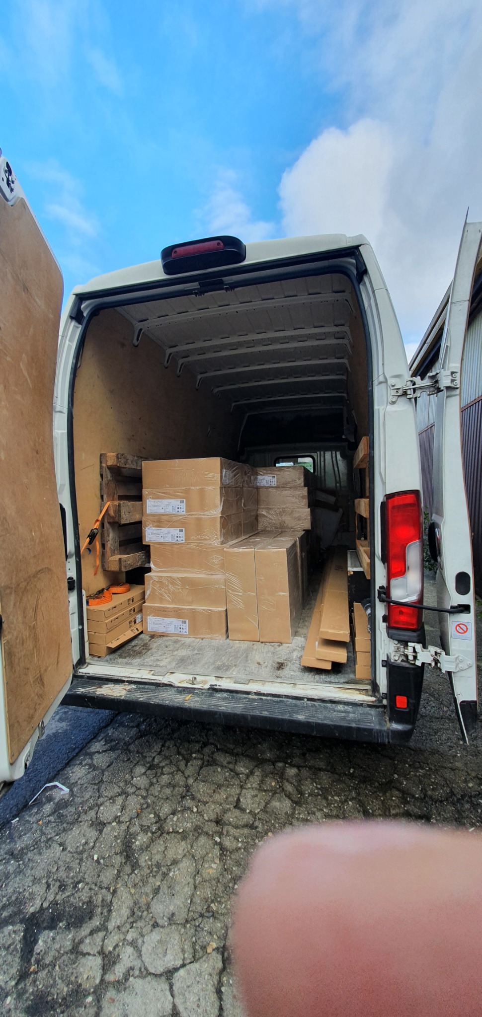 Wnętrze białego busa z otwartymi drzwiami załadowane kartonami zabezpieczonymi folią, drewnianymi listwami i paletami, widok z tyłu pojazdu.