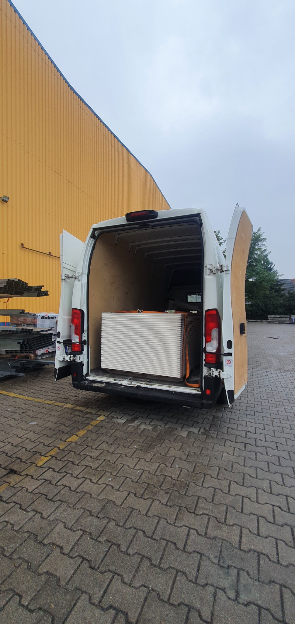 Biały bus z otwartymi drzwiami załadowany płytami gipsowo-kartonowymi zabezpieczonymi pasami transportowymi na tle żółtej ściany magazynu.