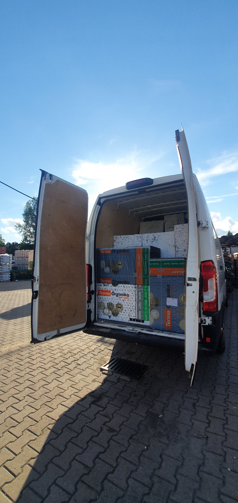 Biały bus z otwartymi drzwiami bagażnika, wypełniony paczkami styropianu termoizolacyjnego 'Termo Organika Galaxy' i 'Termo Organika Silver', widok z tyłu na kostkę brukową i błękitne niebo.