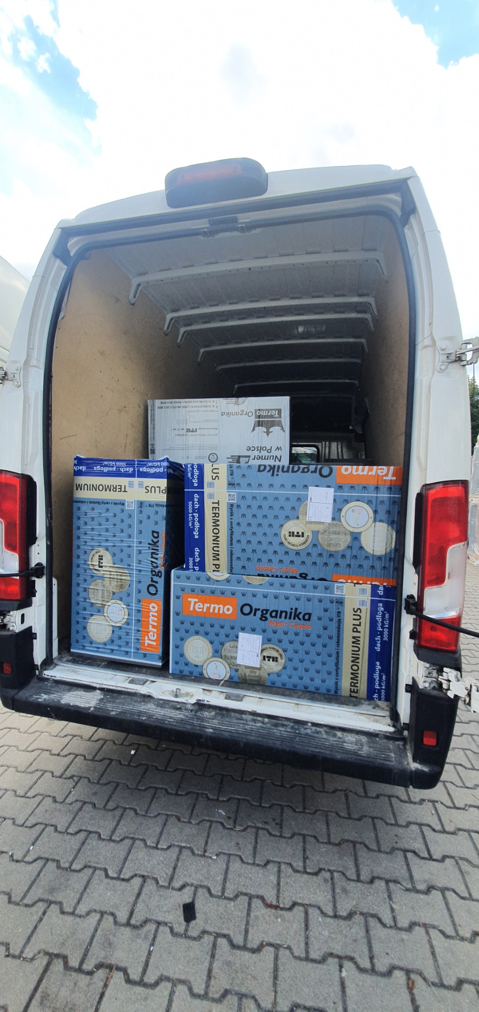 Otwarty bagażnik białego busa wypełniony paczkami styropianu Termo Organika, widok z zewnątrz na kostkę brukową i fragment nieba.