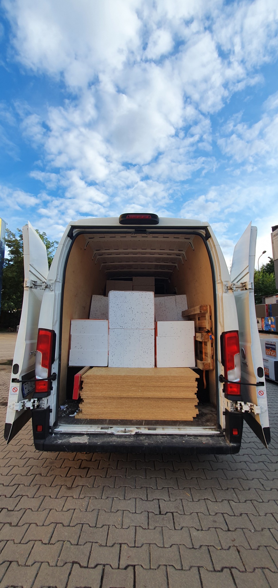 Otwarty bagażnik białego busa załadowany płytami drewnianymi i białymi płytami izolacyjnymi, widok z tyłu na brukowany plac i błękitne niebo z chmurami.