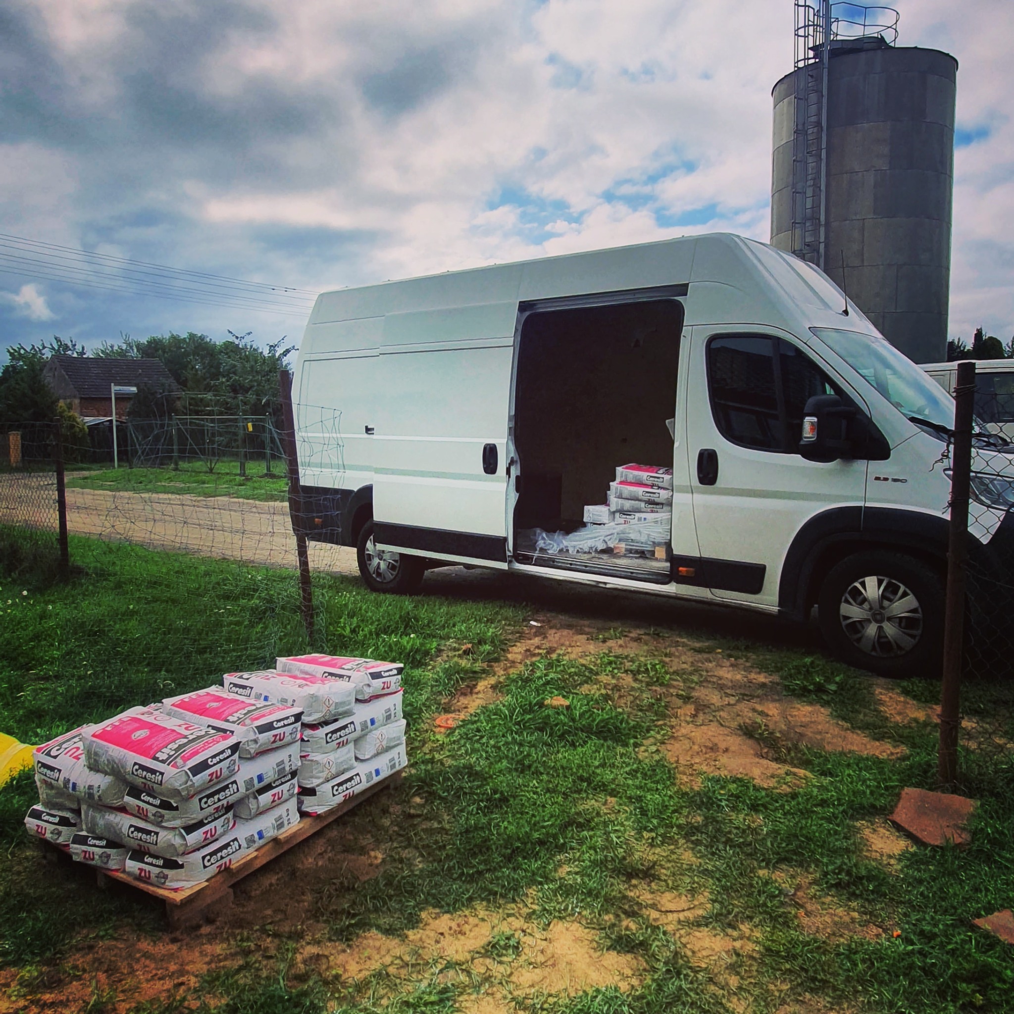 Biały bus z otwartymi drzwiami bocznymi, załadowany workami zaprawy Ceresit ZU, paleta worków przed busem, w tle wieża ciśnień i dom jednorodzinny.