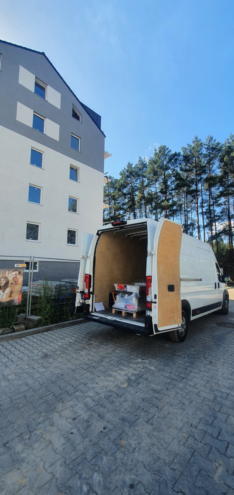 Biały bus z otwartymi tylnymi drzwiami, zaparkowany na brukowanej powierzchni przed nowoczesnym budynkiem mieszkalnym i rzędem wysokich sosen; wewnątrz busa zapakowane na palecie materiały...