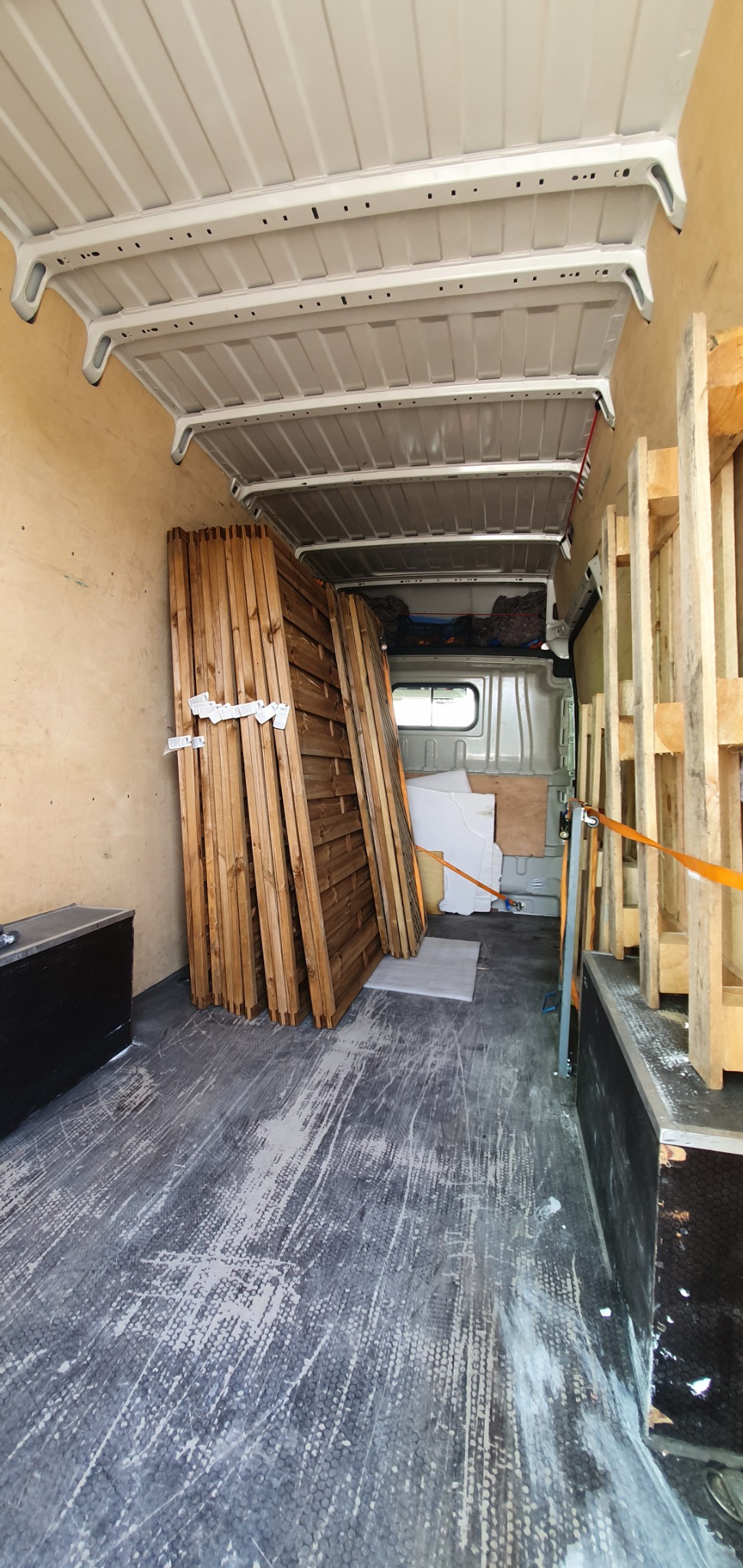 Wnętrze furgonetki załadowane drewnianymi panelami ogrodzeniowymi zabezpieczonymi taśmą, widoczne również palety i inne materiały budowlane.